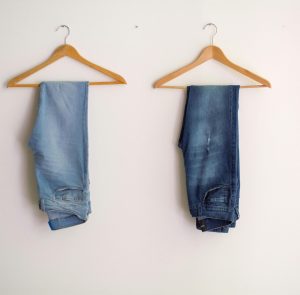Two pairs of blue denim jeans neatly hanging on wooden hangers against a plain wall.